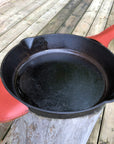 A black cast iron pan with a red leather handle cover fitted on the pan, displayed on a wooden surface.