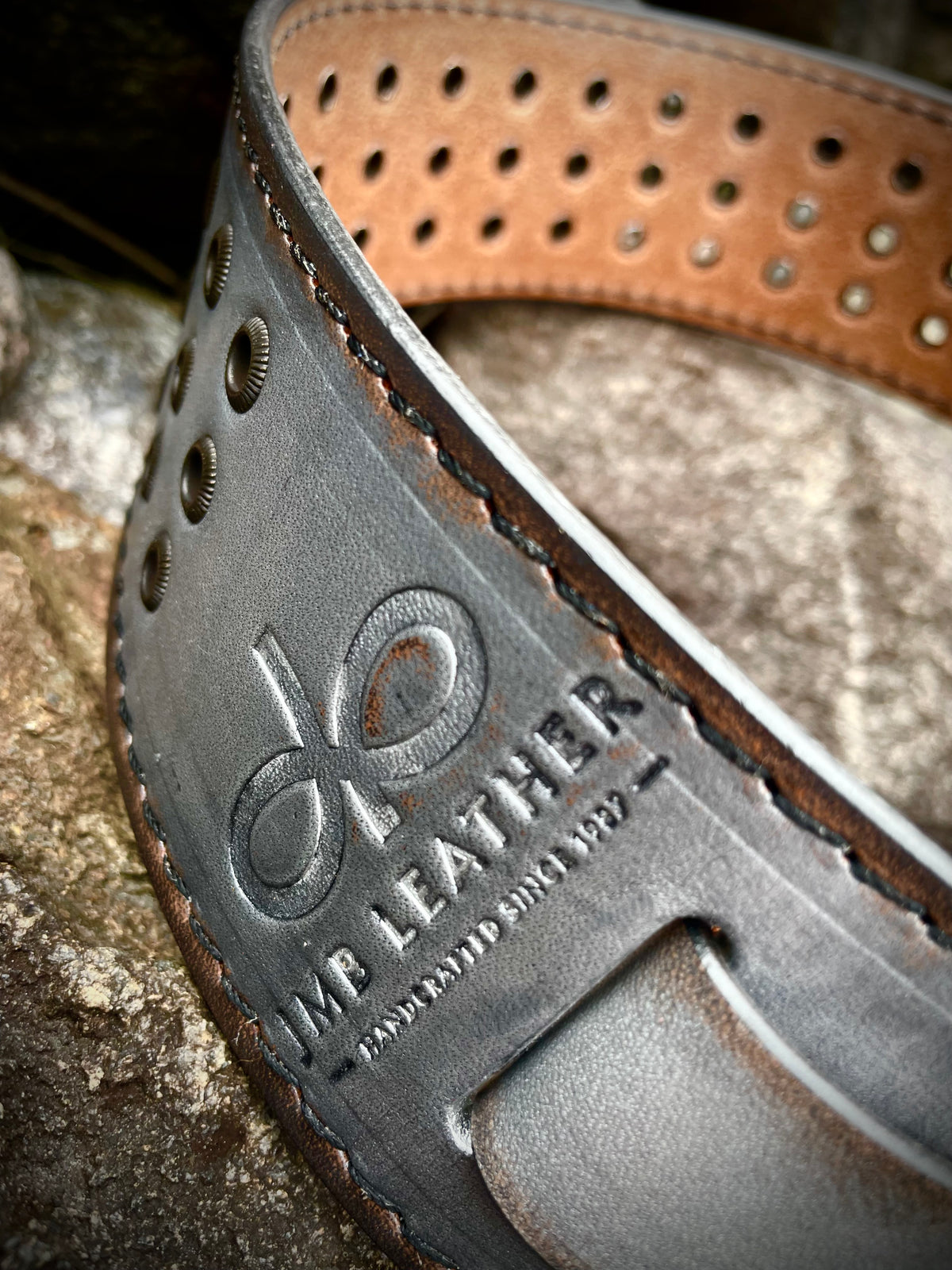 Close-up of a brown leather belt with a silver finish and 'MB Leather' branding on a textured surface.